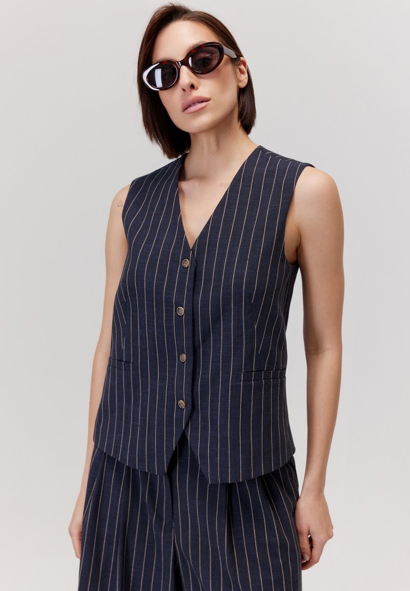 Granatowa kamizelka w pionowe białe paski, dekolt w serek, pięć guzików i kieszenie boczne. W zestawie z dopasowanymi spodniami z plisami.