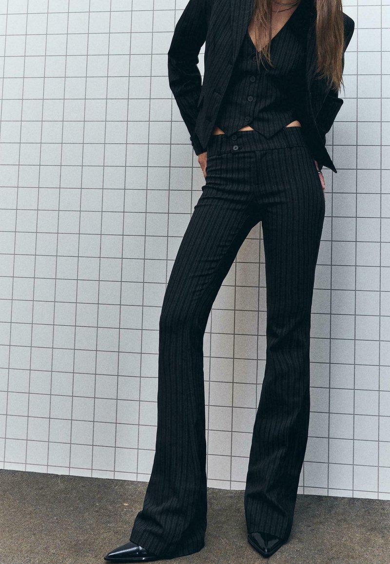 Femme portant un blazer à rayures noires, un gilet assorti et un pantalon évasé, debout contre un mur blanc carrelé, les mains dans le dos.