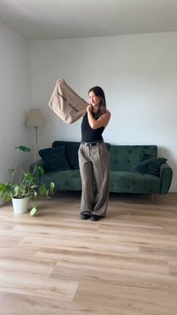 Bēšas tote soma ar melnu tekstu "SAMSØE SAMSØE", valkāta ar augstuma piegriezuma, plašiem pelēkkrāsu biksēm un melnu topu, uz koka grīdas.