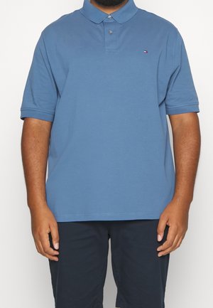 Homme portant un polo à manches courtes bleu avec un petit logo Tommy Hilfiger et un short foncé, debout devant un fond uni.