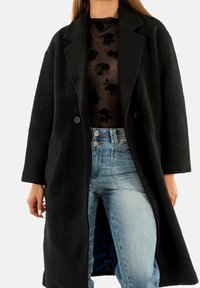 Donna che indossa un lungo cappotto nero a trama, una blusa nera trasparente con stampa floreale e jeans a vita alta di un blu chiaro, su sfondo bianco.