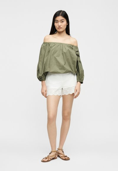 Γυναίκα που στέκεται φορώντας μπλούζα σε στυλ off-shoulder σε ελιά πράσινο, λευκά σορτς με κυματιστή άκρη και καφέ φλατ σανδάλια με λουράκια, σε απλό φόντο.