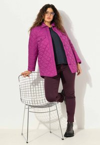 En livfull lila quiltad jacka med knäppning framtill, bärs över en mörk topp och vinröda byxor, matchad med svarta klumpiga stövlar, mot en vit bakgrund.