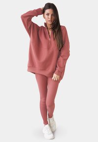 Felpa rosa con colletto con zip, abbinata a leggings fitted coordinati e sneakers bianche. Design minimalista, stile casual.
