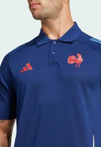 Marineblaues Polohemd mit Kragen, versehen mit einem roten Hahnemanns-Emblem und drei blauen Schulterstreifen. Aus strukturiertem Material gefertigt.