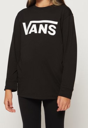 Junge Person trägt ein schwarzes Langarmshirt mit großem weißem Vans-Logo auf der Brust, kombiniert mit schwarzen Hosen.