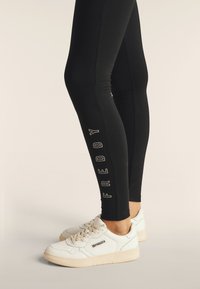 Leggings neri con la scritta "FREDDY" in bianco di lato, abbinati a sneakers bianche con suola testurizzata e logo laterale.