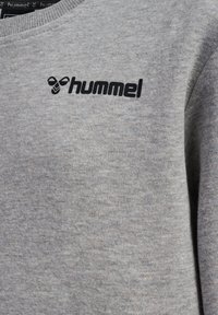 Sweatshirt gris en tissu doux avec un col rond, arborant un logo "hummel" noir sur la poitrine. Design et texture simples.