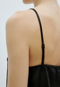 Zwarte satijnen camisole met dunne verstelbare bandjes. Soepele textuur die zachtjes over de rug valt met een geplooide naad aan de onderrand.