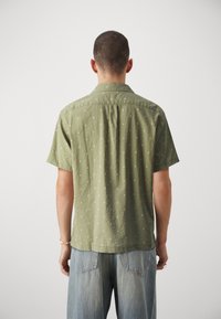 Camicia a maniche corte color verde oliva con pois bianchi, colletto a bottone e una texture morbida. Abbinata a jeans in denim chiaro, vista da dietro.