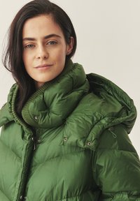 Giacca a vento verde con collo alto, texture trapuntata e bottoni a scatto. Presenta accenti a coste e un cappuccio per maggiore calore.