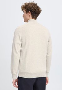 Suéter de cremallera beige claro con cuello y puños acanalados. Presenta una textura suave y un diseño ajustado, adecuado para llevar en capas.
