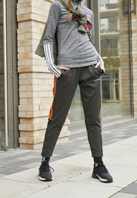 Szary sportowy top z długimi rękawami z białymi paskami. Ciemnozielone spodnie z pomarańczowymi paskami po bokach i kieszeniami zapinanymi na zamek. Czarne trampki.