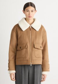 Bruine wollen jas met een witte fleece kraag, twee voorzakken, gouden rits en ribbels aan de mouwen. Eenvoudig, gestructureerd ontwerp.