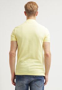 Rumena polo majica kratkih rukavov z gladko teksturo in rebrastim ovratnikom; v kombinaciji z modrimi jeans hlačami, ki prikazujejo sproščen kroj.