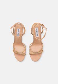 Beige High Heels Sandalen mit überkreuzten Riemen, verziert mit goldfarbenen Verzierungen, aus weichem Wildleder und mit einem Knöchelverschluss.