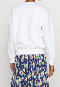 Bílá mikina se žebrovanými manžetami a lemem, která má uvolněný střih a jemnou texturu, spárovaná s barevnou květinovou sukní.