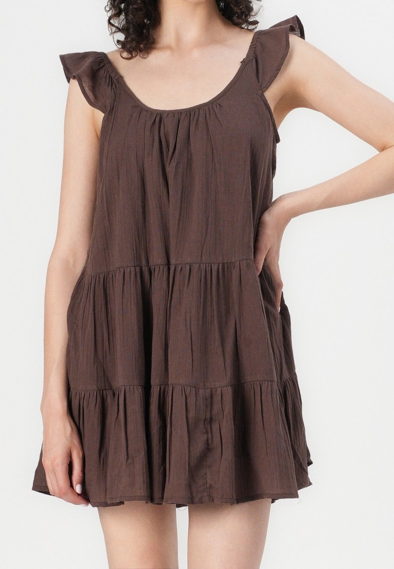 Robe marron sans manches à étages avec bretelles volantées portée par une personne posant une main sur la hanche.