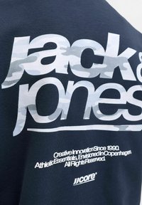 Μπλε πουκάμισο με μεγάλο άσπρο λογότυπο "Jack & Jones" σε καμουφλάζ εκτύπωση και μικρότερο άσπρο κείμενο σχετικά με την καινοτομία της μάρκας και την προέλευση από την Κοπεγχάγη.