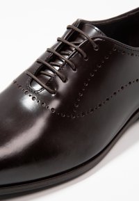 Chaussures en cuir marron foncé avec une texture lisse, présentant des perforations décoratives, un bout arrondi et des lacets marron. Design classique avec une finition brillante.