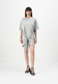 Grey oversized t-shirt dress with a tied knot detail at the waist. Smooth fabric, short sleeves, and a minimalist design. Black high-heeled sandals.