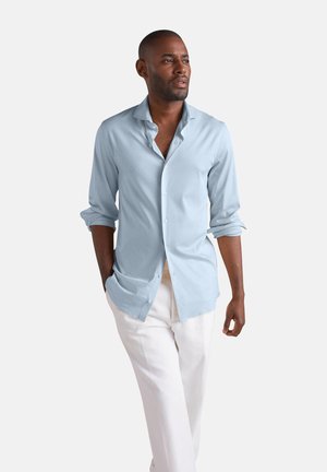Mann geht mit den Händen in den Taschen, trägt ein hellblaues Hemd mit Knöpfen und weiße Hosen vor schlichtem Hintergrund.