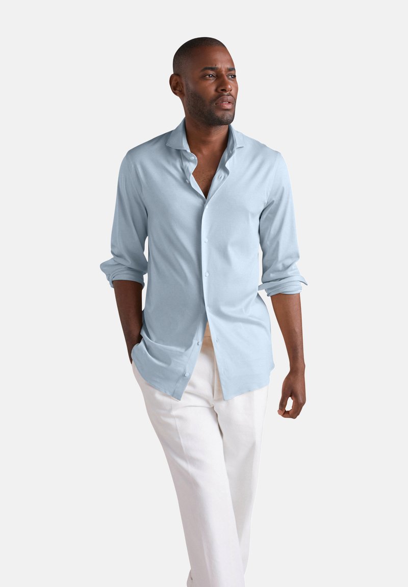 Mann geht mit den Händen in den Taschen, trägt ein hellblaues Hemd mit Knöpfen und weiße Hosen vor schlichtem Hintergrund.