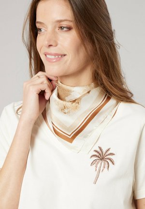 Femme souriante aux longs cheveux bruns, portant une chemise blanche avec une broderie de palmier et une écharpe beige à motifs autour du cou.