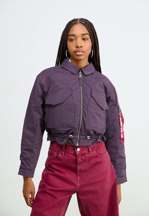 Gekürzte lila Bomberjacke mit zwei vorderen Taschen, Reißverschluss, Kordelzugsaum und seitlichem Branding-Label; kombiniert mit hochtaillierten roten Hosen.