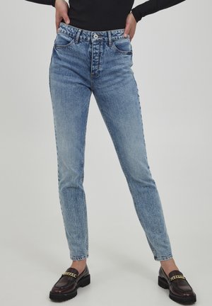 Personne portant un jean droit taille haute bleu clair et des mocassins noirs avec détail chaîne dorée, les mains posées sur les hanches.