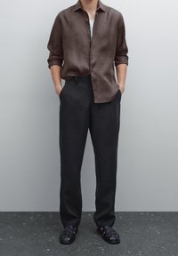 Braunes Leinenhemd, schwarze Hose und schwarze Sandalen. Das Hemd hat eine lockere Passform und Knopfdetails, während die Hose ein flaches Vorderteil hat.