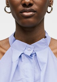 Κοντινό πλάνο ενός ατόμου που φοράει σκουλαρίκια κρίκους και ένα ανοιχτό μπλε αμάνικο τοπ με ψηλό λαιμό και γιακά με κουμπιά.