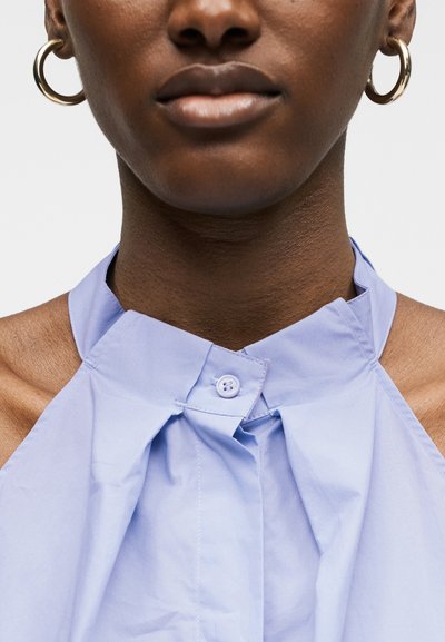Gros plan d'une personne portant des boucles d'oreilles créoles et un haut sans manches à col montant bleu clair avec un col boutonné.