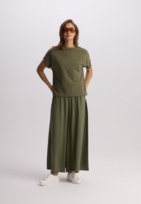 Olive zielona koszulka z podwiniętymi rękawami, zestawiona z oliwkową maxi spódnicą. Model nosi białe sneakersy i okulary przeciwsłoneczne w pomarańczowych odcieniach.