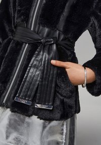 Jachetă neagră din blană sintetică cu accente din piele, fermoar, talie cu curea și textură netedă. Prezintă un logo metalic pe curea.