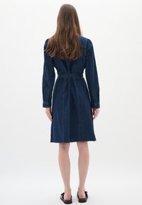 Jeansklänning med långa ärmar, åtsittande midja, knä-längd, mörkblå färg och synliga sömdekorationer på en enfärgad bakgrund.