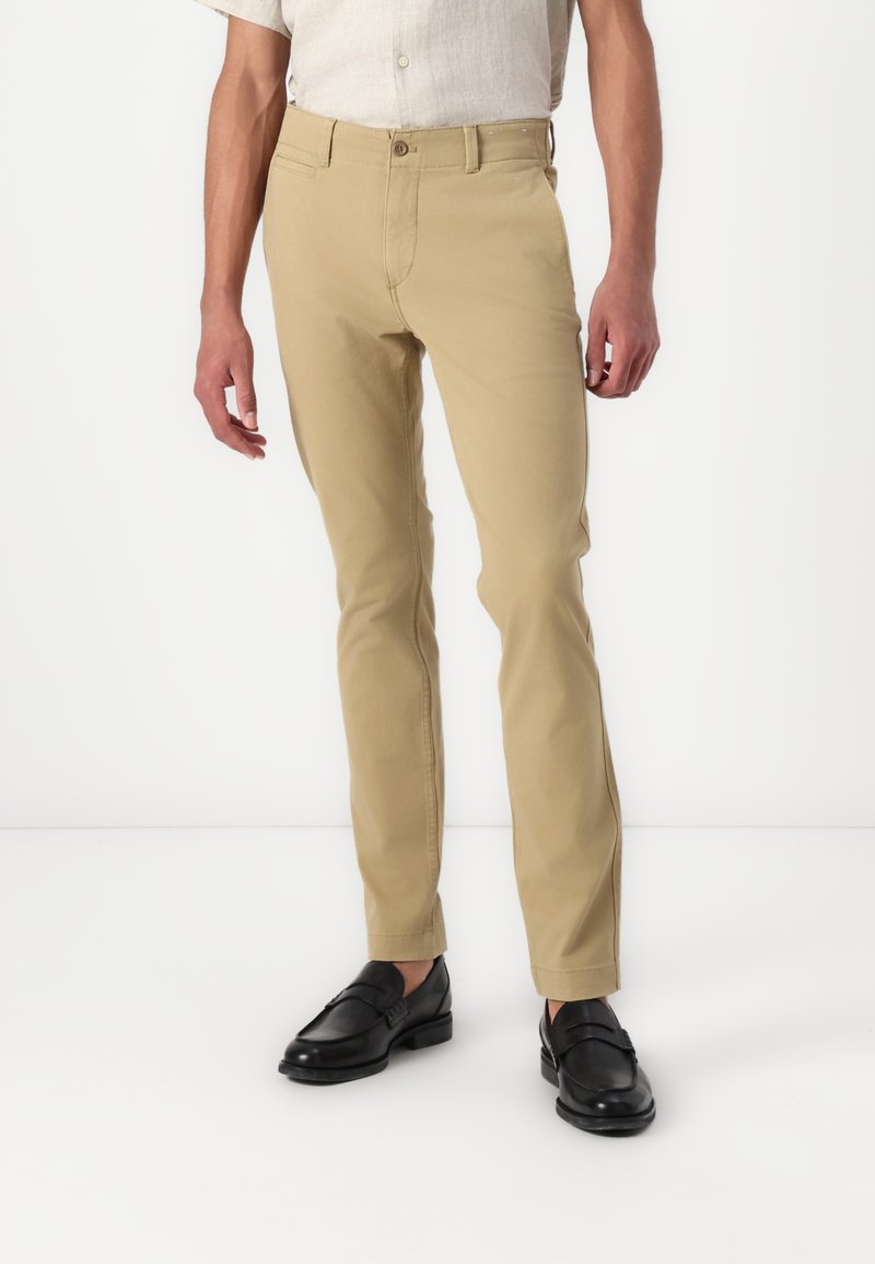 Homme portant un pantalon beige slim, des mocassins noirs en cuir et une chemise claire à manches courtes, debout devant un fond blanc.