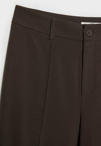 Pantalon marron foncé avec fermeture à bouton, passants pour ceinture et un devant lisse visible en gros plan.