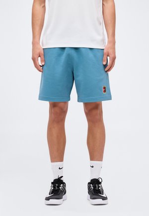 Mężczyzna w turkusowych sportowych szortach, białych skarpetkach z czarnym logo, czarno-białych sneakersach i białej koszulce, stojący na białym tle.