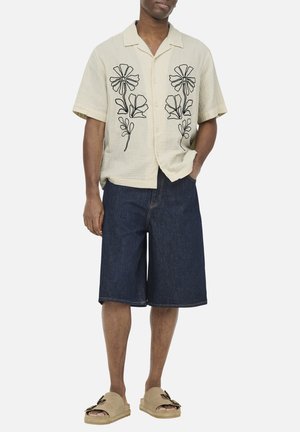 Mann trägt beige kurzärmliges Hemd mit schwarzer Blumenstickerei, dunkle Jeans-Knielange Shorts und beige Sandalen mit Schnallen.
