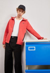 Felpa rossa con zip e logo nero, abbinata a pantaloni larghi neri, accanto a un armadietto blu. Lo sfondo presenta un contesto neutro.