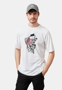 Camiseta de manga corta blanca con un gráfico de un pez que lleva un sombrero azul marino y una camisa de rayas rojas, con acentos de temática náutica.