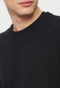 Άνδρας που φοράει μια απλή μαύρη μπλούζα με στρογγυλή λαιμόκοψη, κομμένη κάτω από τη μύτη, δείχνοντας το λαιμό και το πάνω μέρος του στήθους.