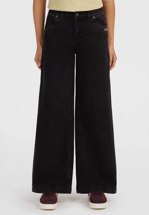 Vrouw draagt zwart hooggetailleerde wijde jeans, beige top en donkerrode sneakers tegen een effen lichte achtergrond.