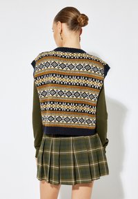 Sweter bez rękawów dzianinowy z wzorami fair isle w granacie, musztardzie i kremie; w połączeniu z zieloną spódnicą w kratę z plisami.