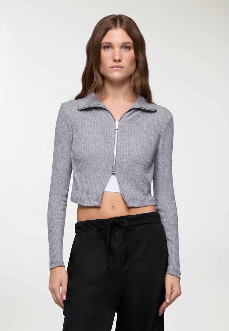 Giacca grigia corta con zip, texture a costine, maniche lunghe e colletto, abbinata a pantaloni neri larghi.