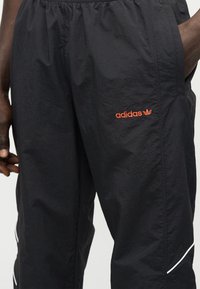 Κοντινό πλάνο μαύρου παντελονιού φόρμας Adidas με πορτοκαλί λογότυπο στο μηρό και λευκή ρίγα κοντά στο τελείωμα, φορεμένο από άτομο με σκούρο χρώμα δέρματος.