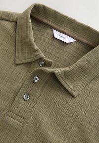Oliven grøn polo t-shirt lavet af tekstureret stof. Har en krave, tre grå knapper og en lille lomme. Brandmærket er synligt indeni.