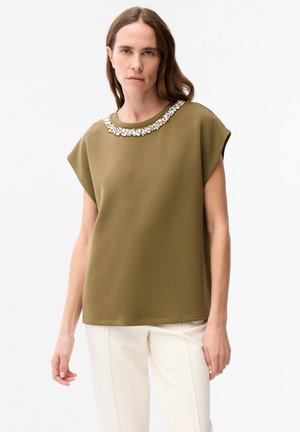 Donna con top verde oliva senza maniche con scollo decorato con perline e pantaloni bianchi, in piedi davanti a uno sfondo chiaro e uniforme.