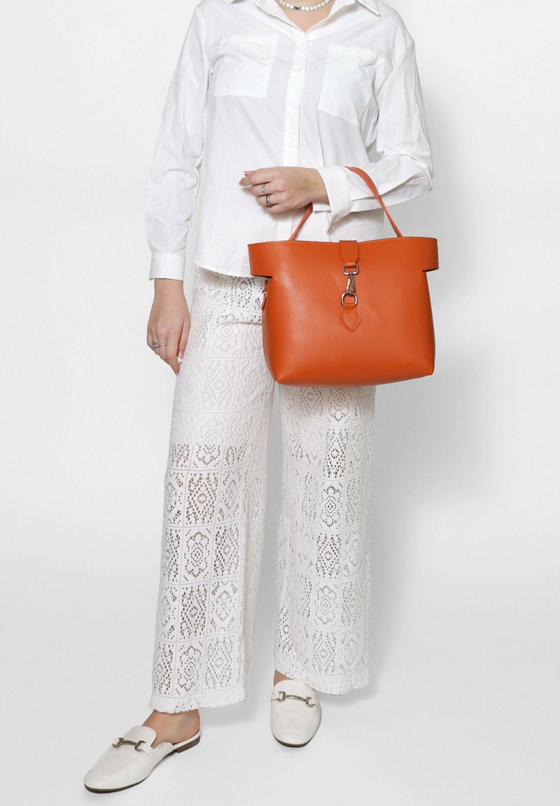Borsa a spalla in pelle arancione con un design strutturato, dotata di un'ampia apertura e ferramenta argentata, abbinata a pantaloni bianchi con motivo a pizzo e scarpe.